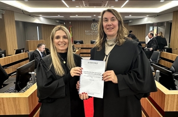Duas mulheres vestindo toga, numa sala de sessão de julgamentos, exibem um documento. Ambas estão sorridentes