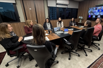 Grupo de sete mulheres e um homem, todos sentados ao redor de uma mesa. Eles observam um telão onde aparecem pessoas em videoconferência