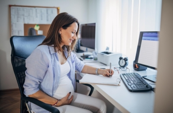 Mulher grávida, sentada, de perfil, trabalhando em um notebook. Ela segura a barriga com um olhar feliz