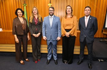 Três mulheres e dois homens em pé posam para foto em um auditório, tendo ao fundo um painel de MDF em tom amarelado