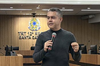 Homem de blusa preta e calça caqui, de pé, fala ao microfone. Ele está em um auditório, tendo ao fundo um brasão do Poder Judiciário iluminado