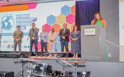 A imagem mostra seis pessoas no palco de um evento, diante de um painel colorido com a inscrição "IV Fórum Internacional de Inovação Social"