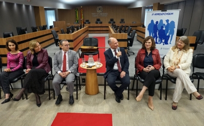 Quatro mulheres e dois homens sentados à frente de um auditório, em cadeiras. Ao fundo, um violão e um banner escrito Aprendizagem