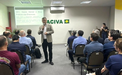 Um homem esta em pé fazendo uma palestra em uma sala de eventos, onde homens e mulheres estão sentados assistindo