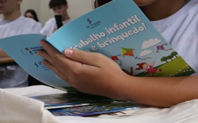 Imagem em close de um jovem segurando uma cartilha com o nome Trabalho infantil não é brinquedo