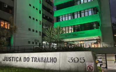 Parte da fachada do TRT-SC iluminada de verde, mostrando três pavimentos. À frente, no chão, um letreiro escrito Justiça do Trabalho