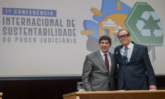 A imagem mostra dois homens de terno posando para uma foto, sorrindo e de frente para a câmera. Eles estão em pé, lado a lado, em frente a um painel que apresenta o título "1ª Conferência Internacional de Sustentabilidade do Poder Judiciário". No fundo, há ilustrações coloridas, incluindo um símbolo de reciclagem e imagens que remetem à natureza e sustentabilidade.