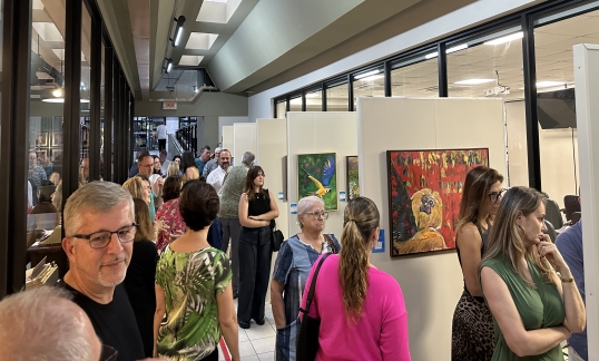 Exposição de arte com quadros coloridos de animais e paisagens, em ambiente moderno, onde um público diverso aprecia as obras.