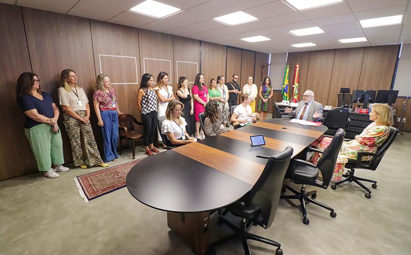 Foto com diversas mulheres em pé, ao lado de uma mesa, com pessoas sentadas à mesa