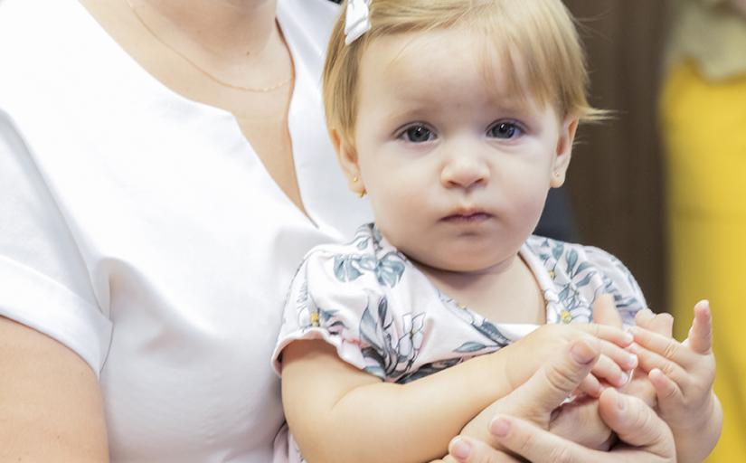 Foto de uma bebê de um ano, loira, no colo da sua mãe e olhando para a foto