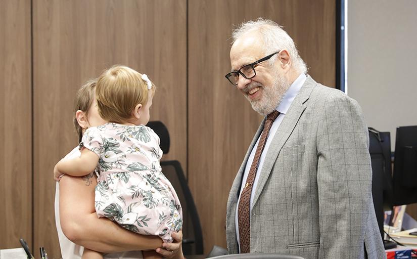 Foto de um homem de barba e cabelo brancos, de óculos de grau, sorrindo para uma bebê do colo da sua mãe