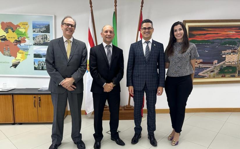 Quatro pessoas, sendo três homens e uma mulher, posam sorrindo em pé para foto com bandeiras ao fundo