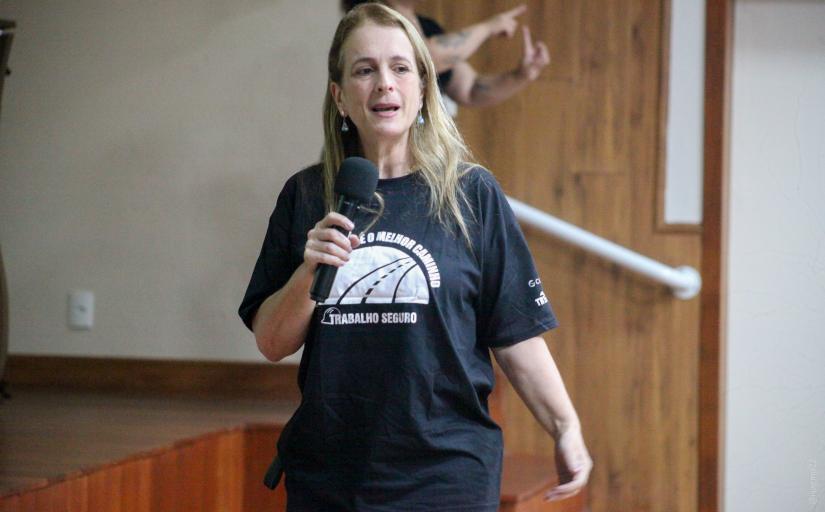 Mulher loira, de cabelos compridos, de camiseta preta, em pé falando ao microfone