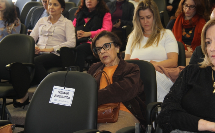 Foto do público que assiste a apresentação, com foco em uma mulher branca de cabelo castanho curto, óculos, blazer marrom e blusa laranja.