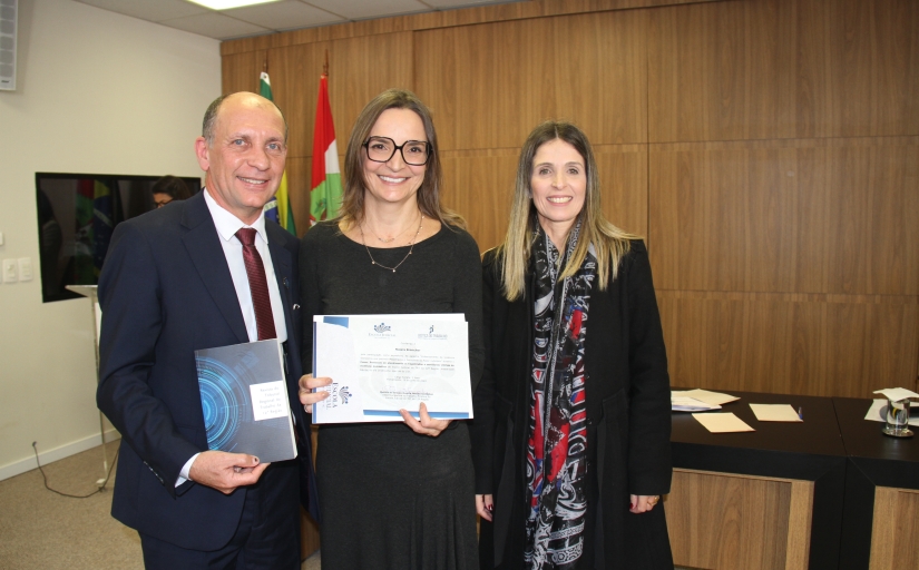 Foto onde está a mesma mulher da imagem anterior segura um certificado e tem ao seu lado esquerdo um homem branco calvo, de cabelo grisalho, com terno azul escuro e gravata vermelha, e à sua direita uma mulher branca loira de cabelo comprido que veste um casaco preto comprido e um xale preto e vermelho.