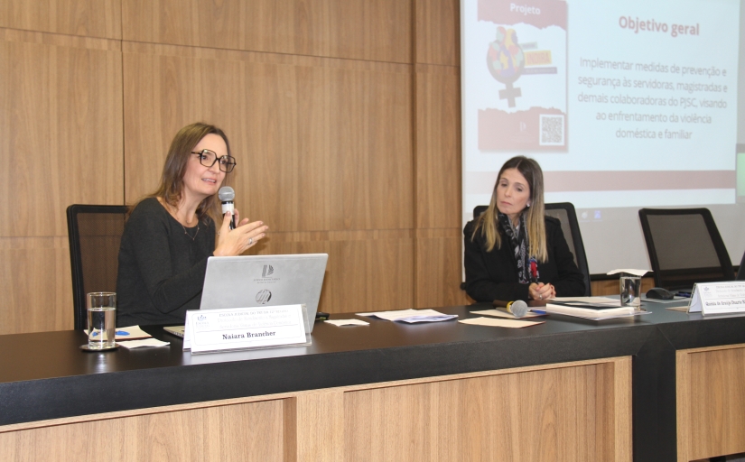 Foto da mesa do auditório onde estão a mulher de óculos de armação preta, que fala ao microfone, e a mulher de xale.