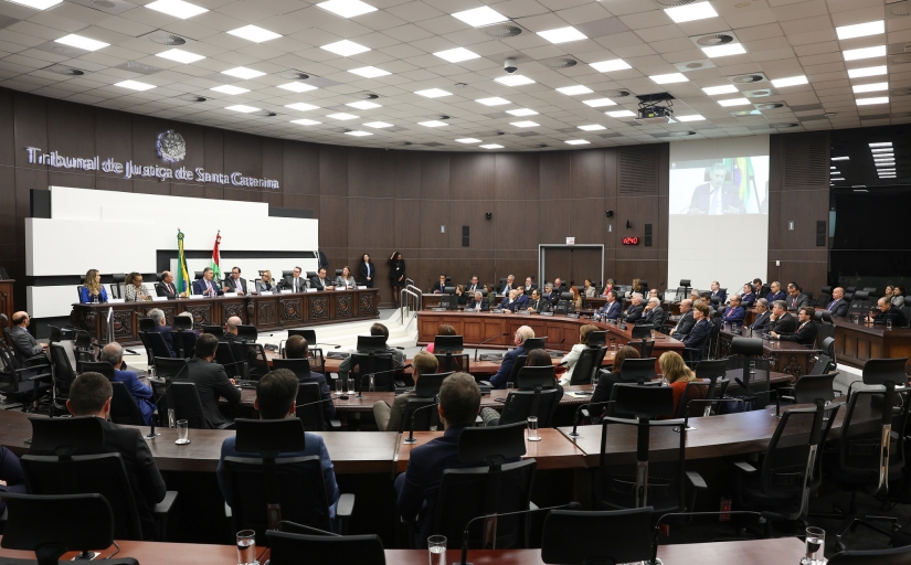 Fotografia de um plenário de Tribunal com mesas em formato semicircular e uma mesa reta à frente, no qual várias pessoas estão sentadas.