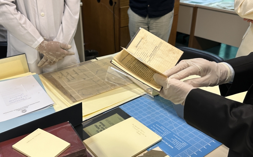 A imagem mostra um par de mãos usando luvas brancas, segurando um livro aberto com páginas amareladas e texto visível. Outro par de mãos com luvas está manuseando uma folha protetora transparente. Há outros documentos e livros na mesa, alguns cobertos com folhas protetoras e outros parcialmente visíveis. O cenário sugere um laboratório, onde documentos antigos estão sendo manuseados, indicando a importância ou fragilidade desses materiais.