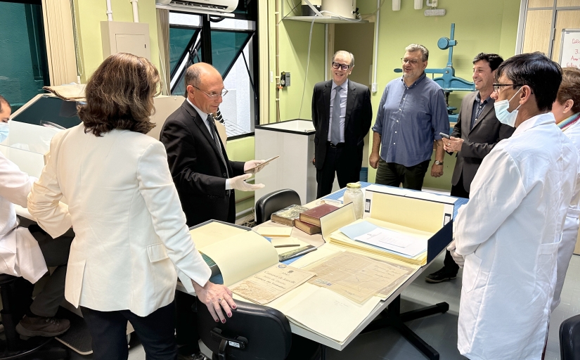 A imagem mostra um grupo de sete pessoas em um ambiente de laboratório. Algumas pessoas estão usando jalecos. Há documentos sobre uma mesa, um quadro branco com anotações e caixas em prateleiras.