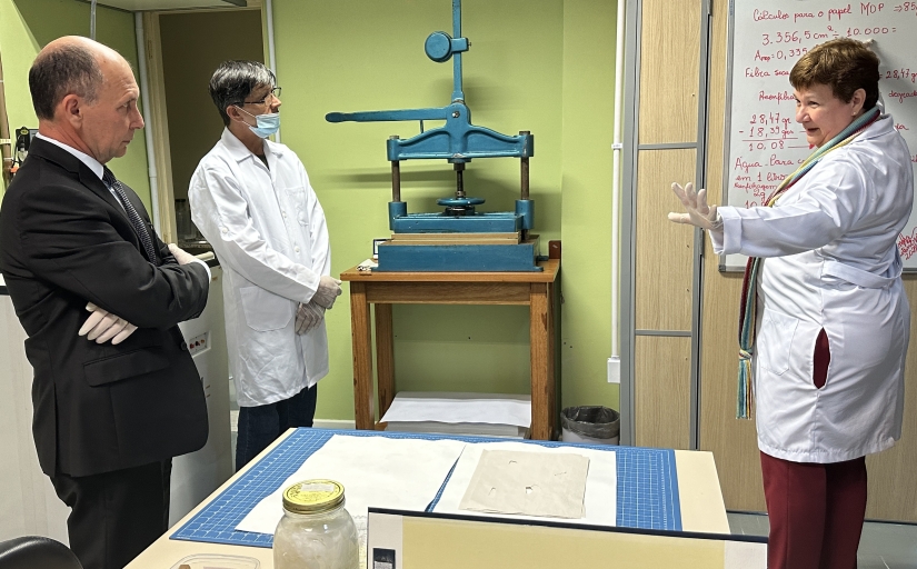 A imagem mostra três pessoas em uma sala com equipamentos de laboratório. A pessoa à esquerda está usando um terno escuro e tem os braços cruzados, olhando para a pessoa no centro, que está usando um jaleco branco. A terceira pessoa, também de jaleco branco, está à direita, perto de uma máquina azul montada em uma mesa. Ao fundo, há um quadro branco com anotações e equações matemáticas.