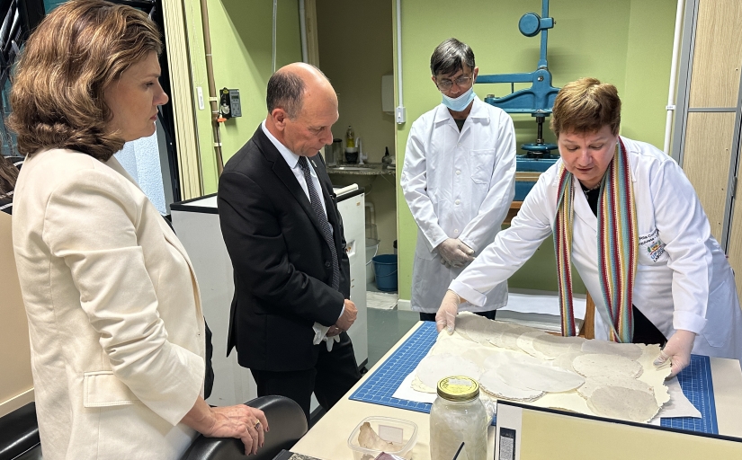 A imagem mostra quatro pessoas em um ambiente que parece ser um laboratório. Uma pessoa está usando um jaleco branco e está atrás de um balcão com papéis espalhados, que parecem estar sendo examinados. Outra pessoa, vestida de forma profissional, está ao lado da pessoa de jaleco. Do outro lado do balcão, há mais duas pessoas que também parecem estar envolvidas na discussão ou exame dos papéis.