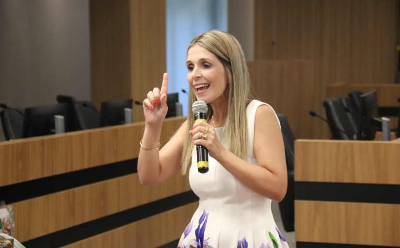 Mulher branca de cabelos loiros compridos, vestindo branco. Ela segura um microfone e aponta para cima.