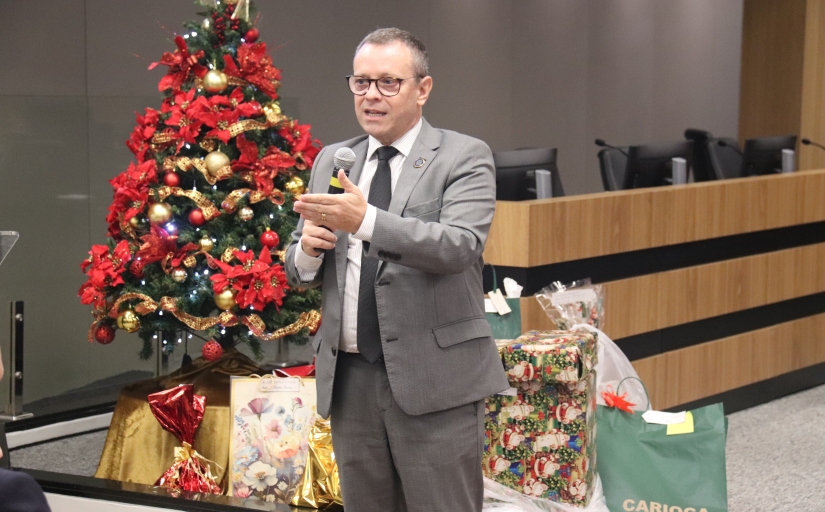 Homem branco de cabelo grisalho curto que veste terno cinza e segura um microfone. Ao fundo um pinheiro de Natal com presentes.