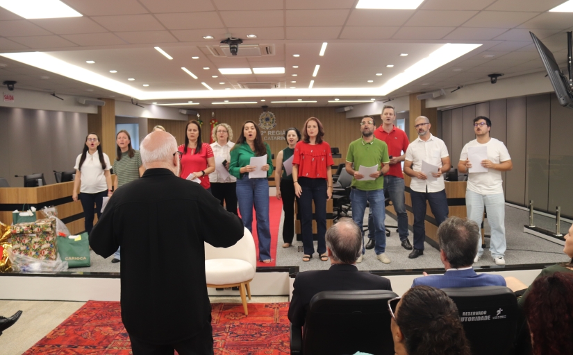 Foto de um coral de doze pessoas vestidas de vermelho, verde e branco, com um homem de terno preto as  conduzindo