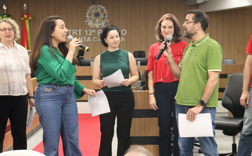 Cinco pessoas do coral, onde um homem e uma mulher estão voltados um para o outro, os dois vestindo verde e segurando um microfone.