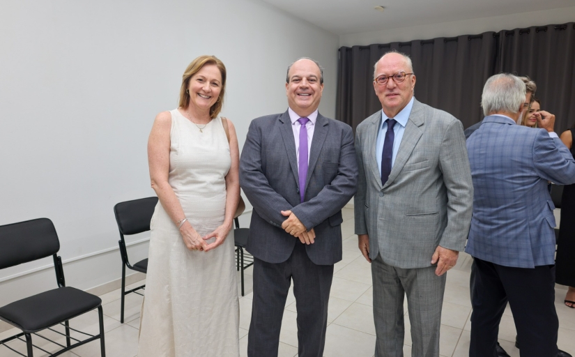 Três pessoas em pé, posando para a foto, sendo uma mulher branca, cabelo curto e loiro, de vestido branco e dois homens brancos, de cabelos grisalhos, vestindo terno