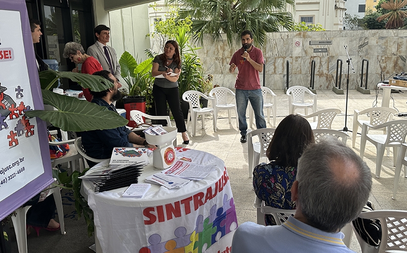 Homem de camiseta vinho falando ao microfone para diversas pessoas sentadas