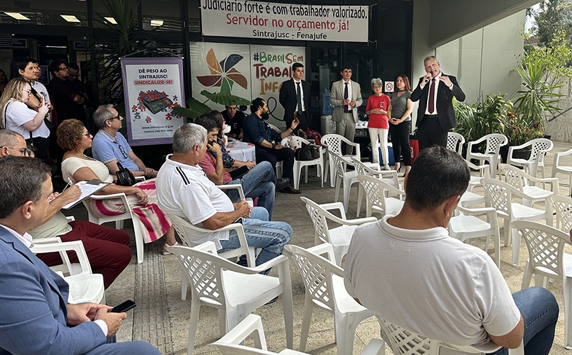 Homem de terno preto e gravata falando ao microfone para diversas pessoas sentadas.