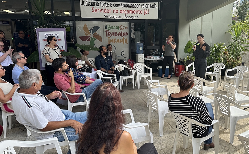 Mulher vestindo calça e camisa pretas falando ao microfone para diversas pessoas sentadas.