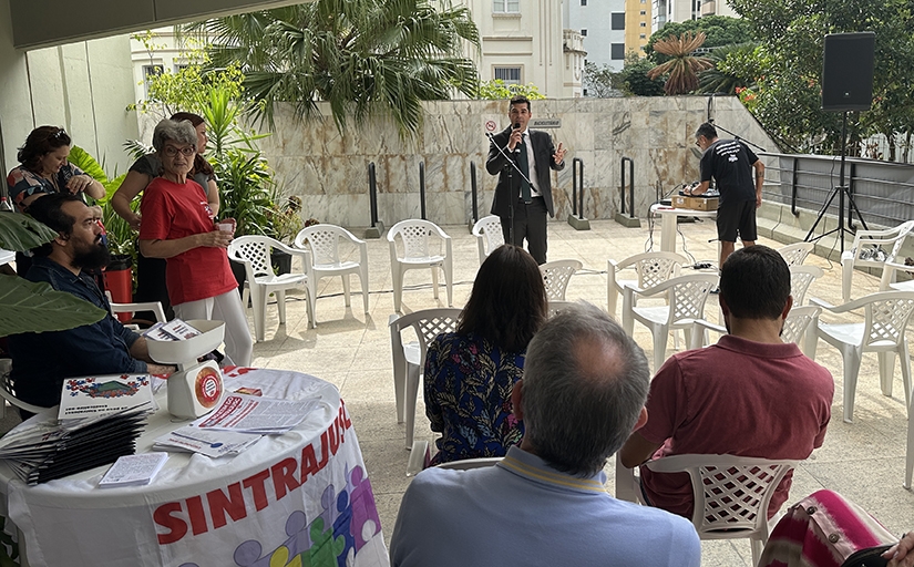 Homem de terno cinza e gravata falando ao microfone para diversas pessoas sentadas.