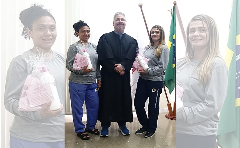 Homem trajando toga posa entregando brindes para duas moças alusivos ao Outubro Rosa