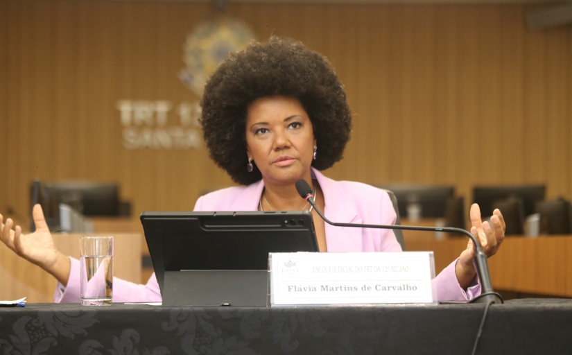 Mulher negra, de cabelo black power, dá palestra. Ela está sentada e com os braços abertos