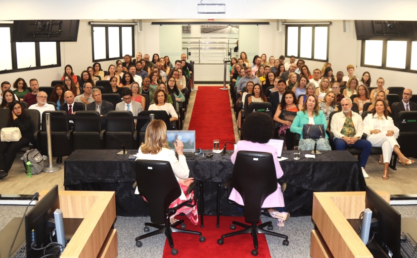 Foto aberta mostra palestrantes de costas e público de frente durante um evento