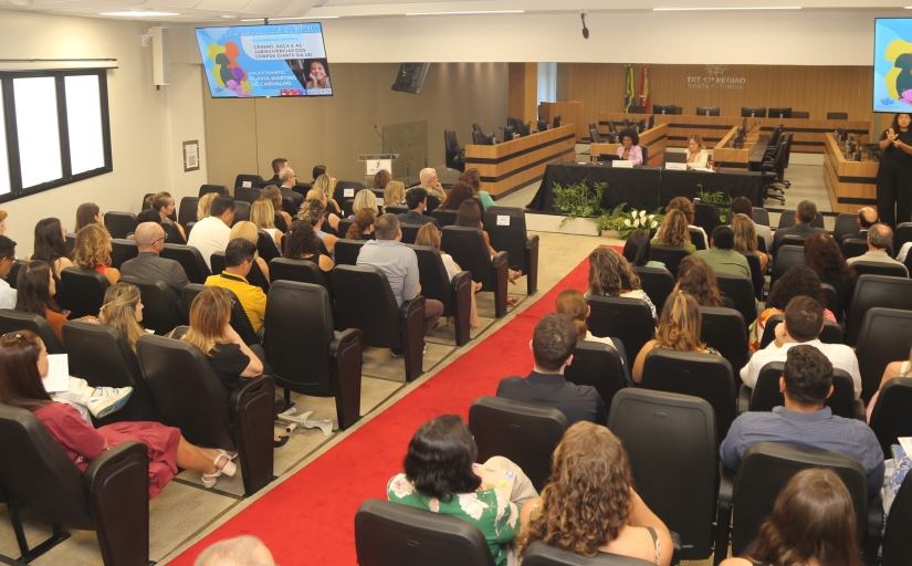 Foto tirada do fundo de um auditório mostra cerca de 100 pessoas e a mesa do evento