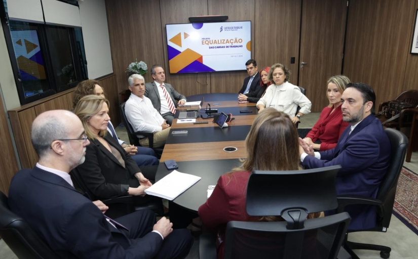 Cinco homens e seis mulheres estão sentados em uma mesa oval de reunião. No fundo da sala tem um telão escrito equalização