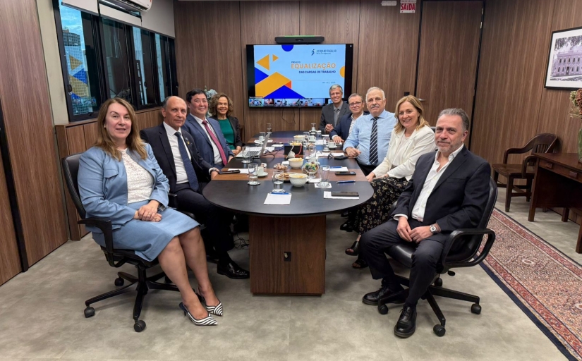 Seis homens e três mulheres estão sentados em uma mesa oval de reunião. No fundo da sala um telão com o slide escrito equalização
