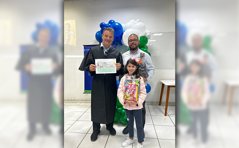 Homem de toga e outro homem posam com menina segurando prêmio em evento com balões ao fundo