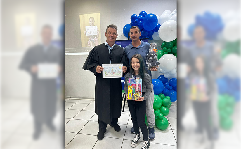 Homem de toga e outro homem posam com menina segurando prêmio em evento com balões ao fundo