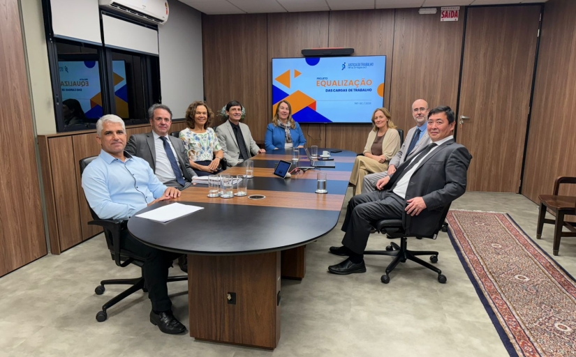 Cinco homens e três mulheres estão sentados em uma mesa oval de reunião. No fundo da sala um telão com o slide escrito equalização