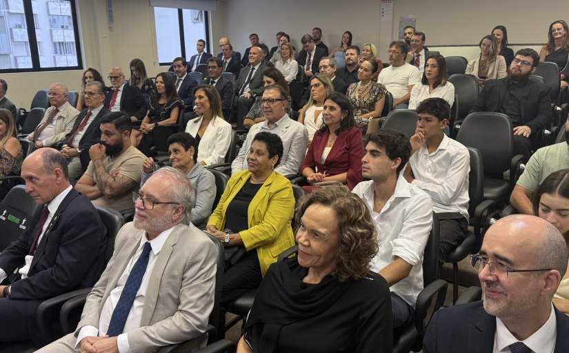 Cerca de 60 pessoas sentadas em um auditório observam uma pessoa falando